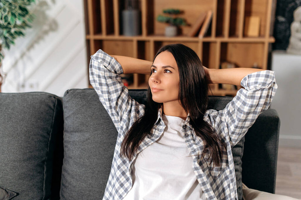 Relaxed happy pretty, caucasian brunette woman, put hands behind head, sits in modern living room on comfortable sofa, enjoy calmness and alone at home in a weekend, looks to the side, dreaming, smile - Foto, imagen