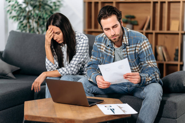 Sad caucasian couple sitting on the couch, the guy and the girl are working from home together, use a laptop, look at graphs, are unhappy with the results, low profit, thinking about strategy - Foto, immagini