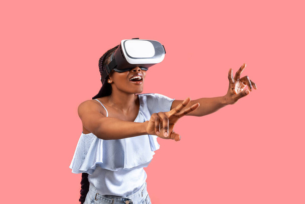 Eccitato giovane donna nera in VR occhiali interagire con il cyberspazio, giocare online in realtà virtuale su sfondo rosa studio. Donna afroamericana emotiva che utilizza tecnologie moderne - Foto, immagini