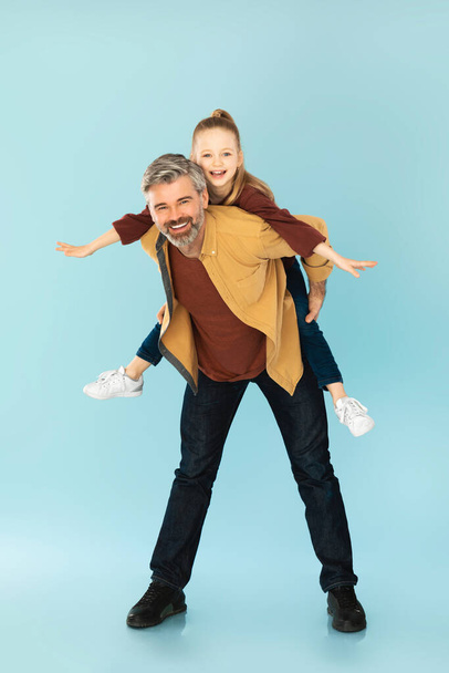 Happy Middle Aged Daddy Carrying His Kid Daughter Piggyback Posing And Having Fun Standing Over Blue Studio Background. Family Concept. Full Length, Vertical Shot - Photo, image