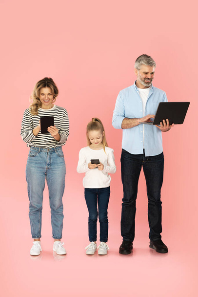 Happy Family Using Gadgets Browsing Internet On Smartphone, Laptop And Digital Tablet Computers Standing Together Over Pink Studio Background. Vertical Shot - Foto, Imagem