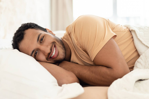 Fröhlicher arabischer Typ, der lächelnd in die Kamera im Bett liegt, den Kopf auf dem Kopfkissen liegend posiert und es sich im modernen Schlafzimmer zu Hause gemütlich macht. Erholung und häuslicher Komfort - Foto, Bild