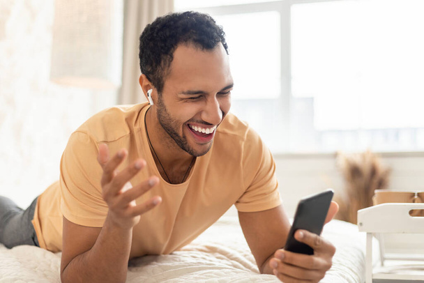 Счастливый ближневосточный мужчина слушает музыку на сотовом телефоне с натянутыми наушниками, весело лежа на кровати дома. "Millennial Guy Using Musical Application Enjoying New Song". Селективный фокус - Фото, изображение
