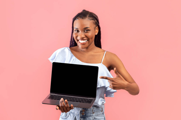 Neşeli genç siyahi bayan pembe stüdyo arka planında boş ekranla dizüstü bilgisayarı işaret ediyor. Afro-Amerikalı kadın reklam için taşınabilir bilgisayarını gösteriyor. - Fotoğraf, Görsel