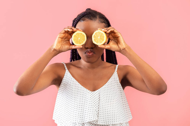 Fiatal fekete nő fonatokkal, citromféléket tartva a szemei előtt, ajkakat csörgetve rózsaszín stúdió háttérrel. Millenniumi afro-amerikai hölgy citrusféléket tart az arca mellett. Nyári méregtelenítés - Fotó, kép