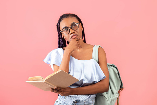 Vollbusige schwarze Studentin mit Rucksack, aufgeschlagenem Lehrbuch, Aufgabenüberlegung, Prüfungsvorbereitung auf rosa Studiohintergrund. Nachdenkliche junge Frau bereitet sich auf Schultest vor - Foto, Bild