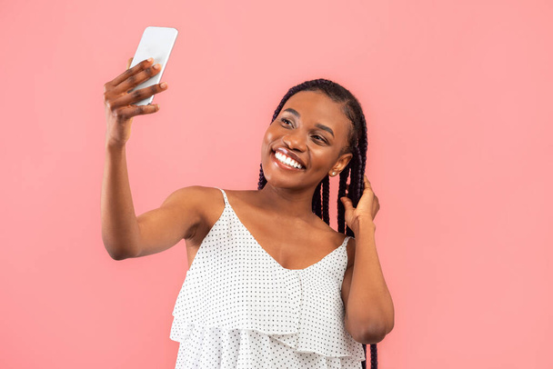 Очаровательная молодая чернокожая женщина делает селфи со смартфоном, снимая контент для своего видеоблога на розовом фоне студии. Веселая афроамериканка чатится онлайн с другом, фотографирует себя - Фото, изображение