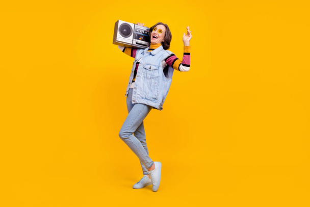 Full body photo of sweet grey hairdo old lady hold boom box wear eyewear sweater vest jeans shoes isolated on yellow color background. - 写真・画像