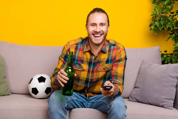 Photo of cheerful guy sit sofa hold remote control drink beer watch tv wear plaid shirt isolated yellow color background. - 写真・画像