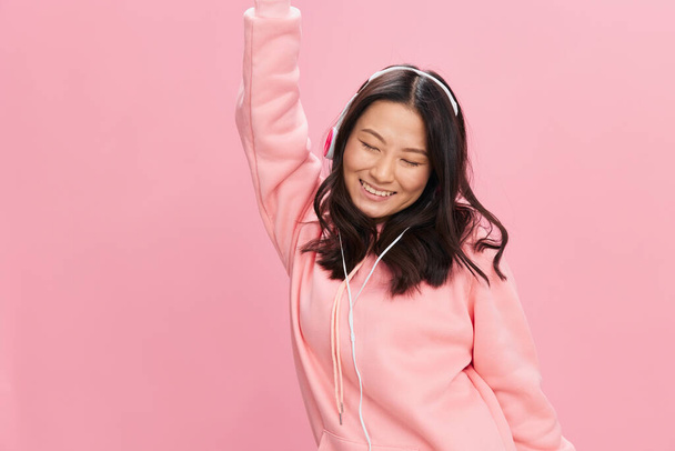 Overjoyed Asian student young lady in pink hoodie sweatshirt with cute headphones dance moving to music posing isolated on over pink studio background. Good offer. Sound streaming platform ad concept - Foto, immagini