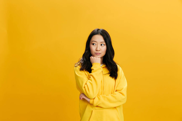 Хм. Удумлива стомлююча азіатська студентка-молода жінка в жовтому капюшоні сидить на кулаці Подумайте про проблеми освіти, що позують ізольовані на помаранчевому студійному фоні. Крута пропозиція. концепції емоцій людей - Фото, зображення