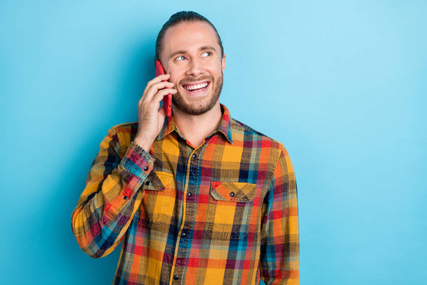 Foto von beeindruckt junge Schwanz Frisur Kerl sprechen Telefon suchen leeren Raum tragen kariertes Hemd isoliert auf blauem Hintergrund. - Foto, Bild