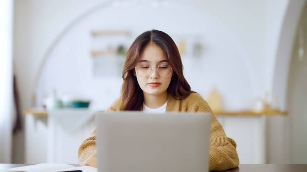 Een glimlachende Aziatische jonge vrouw die thuis op laptop werkt. Jonge Aziatische student met behulp van computer afstandsonderwijs, virtuele opleiding, e-learning, kijken naar online onderwijs webinar in huis - Foto, afbeelding