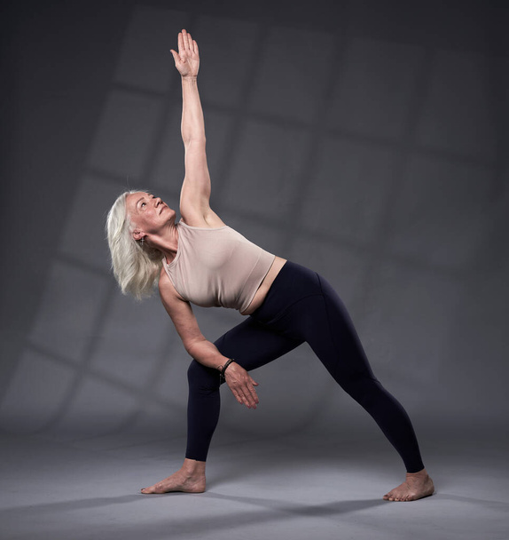 Mujer mayor con entrenamiento de pelo blanco yoga, plano de estudio sobre fondo gris - Foto, imagen