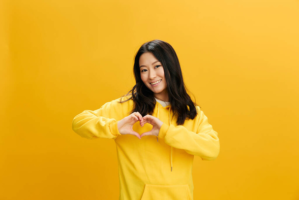 Bella innamorarsi felice studentessa asiatica giovane donna in felpa gialla felpa mostrano segno a forma di cuore posa isolata su sfondo arancio studio. Bella offerta. Persone Emozioni concetto - Foto, immagini