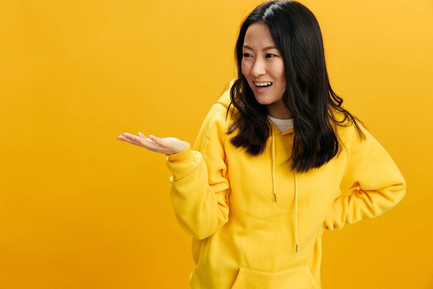 Hey, schau dir das an. Aufgeregt lächelnd hält eine junge Asiatin in gelbem Kapuzenpullover unsichtbare Gegenstände in der Hand, die isoliert auf orangefarbenem Studiohintergrund posieren. Cooles Angebot. Menschen-Emotionen-Konzept - Foto, Bild