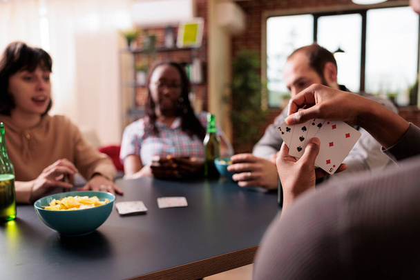 Selective focus of african american man holding cards while playing with best friends at home. Happy multiethnic people sitting at table in living room while enjoying fun leisure activity together. - Photo, Image