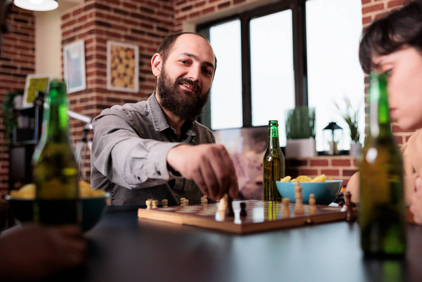 Lächelnder Mann bewegt Schachfigur an Bord, während er mit engen Freunden am Tisch sitzt. Multiethnische Menschen im heimischen Wohnzimmer spielen Brettspiele, während sie Snacks und Getränke konsumieren. - Foto, Bild