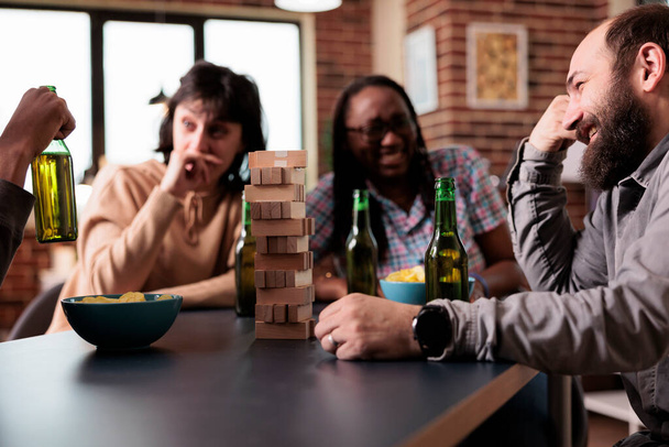 Amigos felices multiétnicos sentados a la mesa en la sala de estar mientras juegan juegos de sociedad juntos. Diversas personas se divierten disfrutando de aperitivos, cerveza y divertidas actividades de ocio en casa. - Foto, Imagen