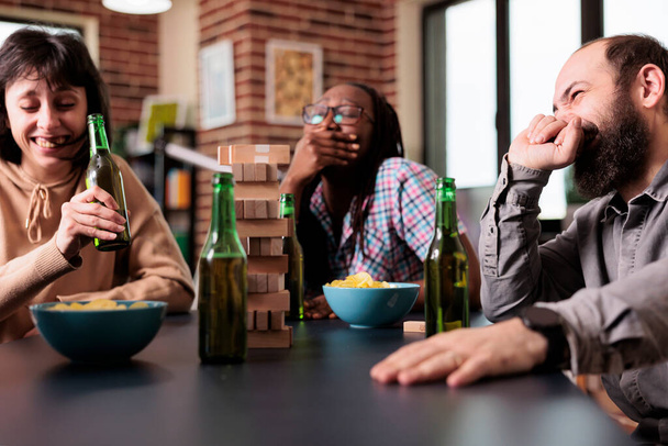Multikulturní skupina přátel se srdečně směje, když spolu doma hrají společenské hry. Šťastný veselý rozmanité lidi sedí u stolu v obývacím pokoji, zatímco hraje s dřevěnými bloky. - Fotografie, Obrázek