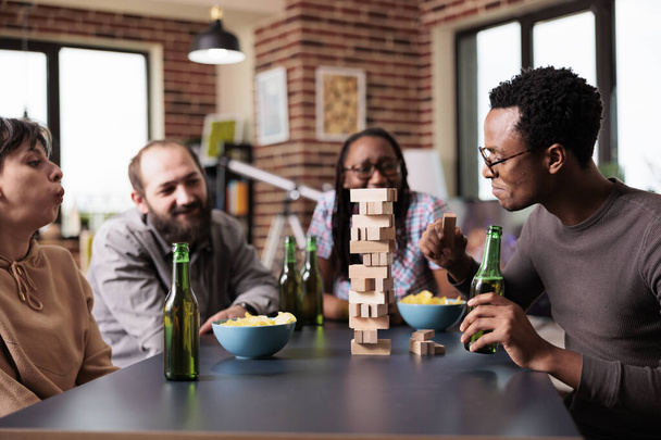 Wieloetniczna grupa przyjaciół siedzi przy stole w salonie, grając razem w gry towarzyskie. Różnorodne osoby siedzące w domu, ciesząc się grą party z drewnianymi klockami i konstrukcji wieży. - Zdjęcie, obraz