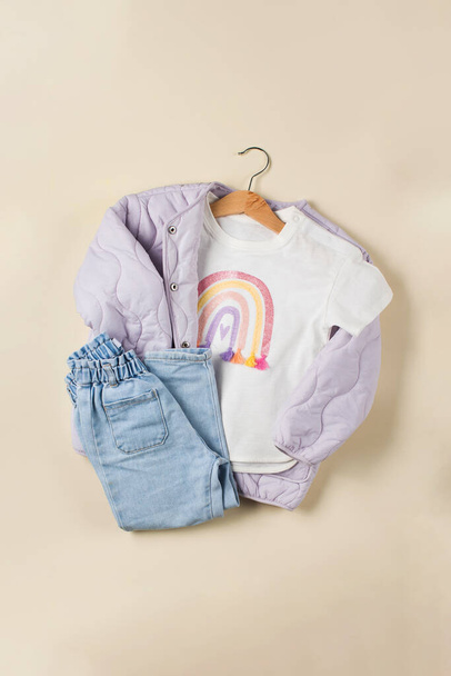 Chaqueta para niños lila, jeans y camiseta blanca con un arco iris en una percha sobre un fondo beige. Imagen de moda para una chica. Puesta plana. - Foto, imagen