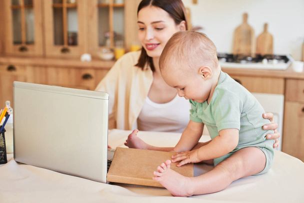 A young Caucasian pretty woman, a mother, works at home at a laptop while her little child sits nearby on the table. Working mom concept. - Foto, Imagem