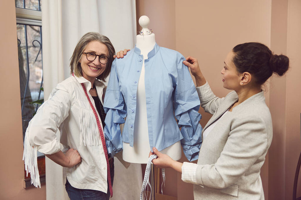 Старший дизайнер моди посміхається позує з моделлю дресмейкера в синій сорочці з нової колекції, в той час як її колега робить вимірювання і працює над поліпшенням деталей нового одягу
 - Фото, зображення