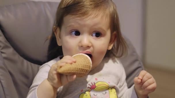 赤ちゃんの女の子はコーンからアイスクリームを食べ、自宅で、屋内で高い椅子に座っている。家族の概念。甘いデザート。休日のコンセプト. - 映像、動画