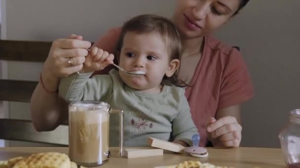 La mamma fa colazione con il suo bambino tenendola tra le braccia e nutrendola con un cucchiaio, dando schiuma di cappuccino al latte. Bella mamma che alimenta il bambino, grande - Filmati, video