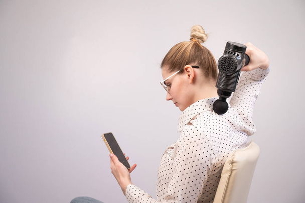 A young woman sits on a chair, uses a smartphone and makes herself massage with an electro massage gun - Fotoğraf, Görsel