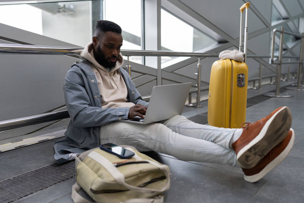 Pensive Afrikkalainen Amerikan mies opiskelija matkustava keltainen matkalaukku keskittyy koulutukseen verkossa, kannettavan tietokoneen, kommunikoida sosiaalisessa mediassa, valmistautuu tentti tai testi istuessaan lentokentällä. - Valokuva, kuva