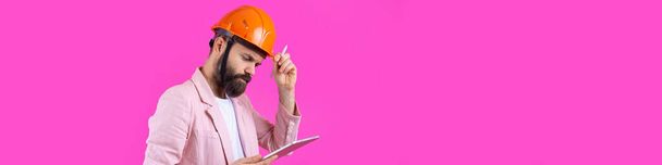 Porträt eines jungen Bauingenieurs mit orangefarbenem Hut, in einem rosafarbenen Sakko auf rotem Studiohintergrund. Ein Mann mit einer Tablette in der Hand. - Foto, Bild