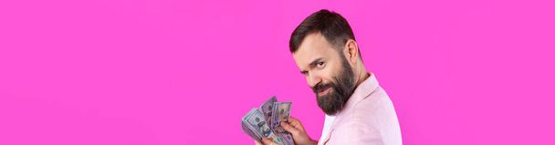 Πορτρέτο ενός ικανοποιημένου νεαρού επιχειρηματία με γενειάδα ντυμένου με ροζ σακάκι που μας δείχνει χαρτονομίσματα δολαρίου σε φόντο κόκκινου στούντιο. Γεύση, μυρωδιά του χρήματος. - Φωτογραφία, εικόνα
