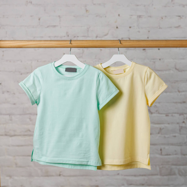 blue and yellow children's T-shirts hang on a hanger against a white brick wall. design blank - Fotografie, Obrázek