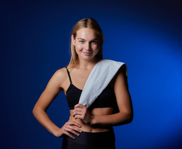 Image of a caucasian blonde woman in a tracksuit smiling with a towel, against a dark blue wall background - Fotó, kép