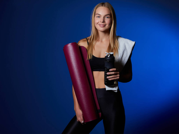 Smiling fitness woman in top and leggings takes break after training, holds bottle of water, wipes sweat with towel, being energetic runner or jogger, feels thirsty. People, wellness, vitality concept - Photo, Image