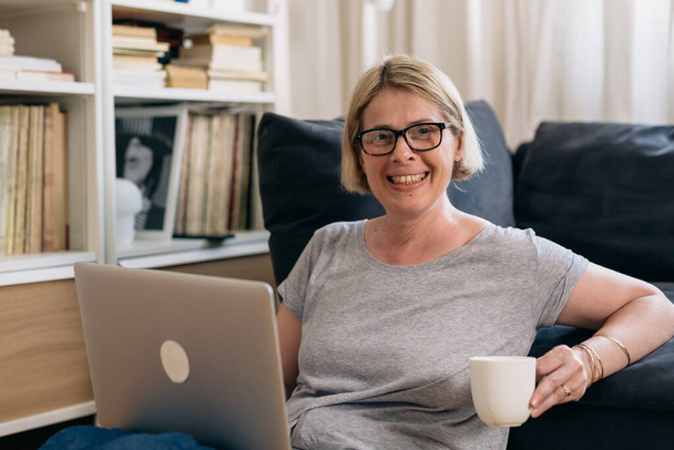 caucasian adult blonde hair woman with eyeglasses holding cup of coffee, sitting in her living room and looking at camera - 写真・画像