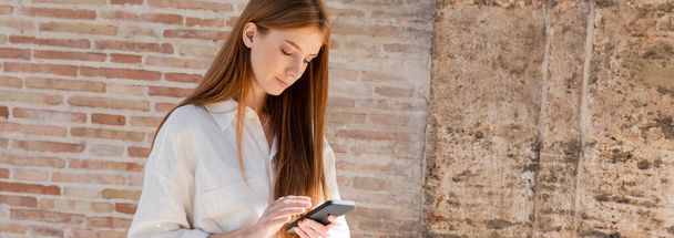 young redhead woman using smartphone on european street near brick wall, banner - Photo, image