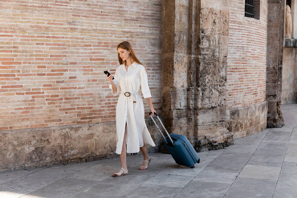 full length of redhead woman walking with baggage and using smartphone on european street - Photo, Image
