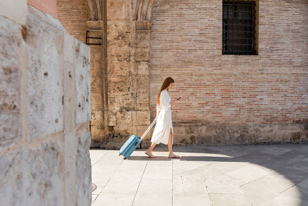 side view of redhead woman walking with baggage and using smartphone on european street - Φωτογραφία, εικόνα