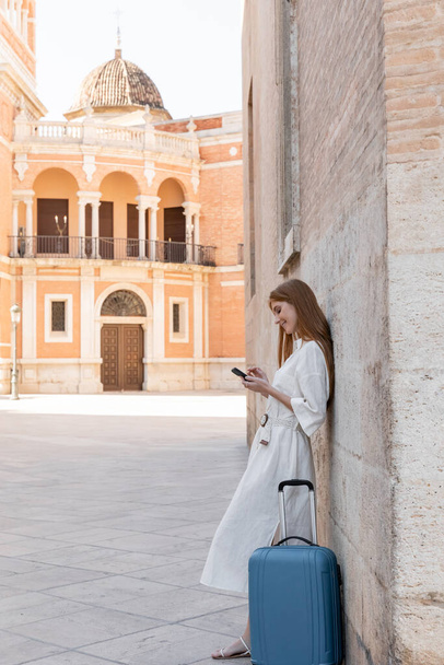 täyspitkä iloinen punapää nainen seisoo lähellä matkalaukkua ja käyttää älypuhelinta kadulla Valencia - Valokuva, kuva