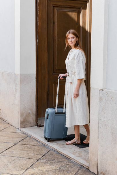 повна довжина рудої жінки в білій сукні тримає ручку багажу і стоїть біля дверей на вулиці у Валансії
 - Фото, зображення