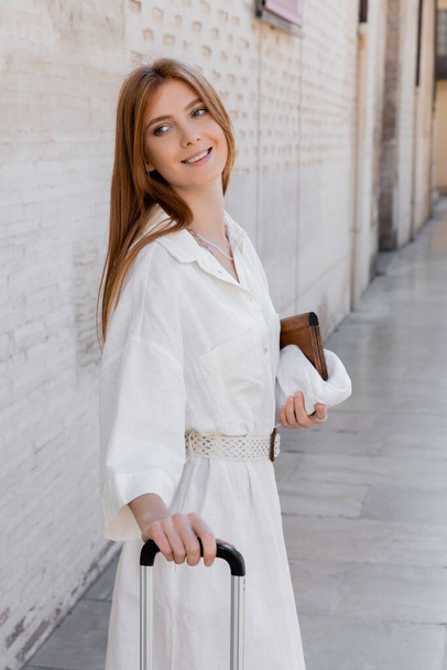 χαρούμενη γυναίκα με φόρεμα που κρατάει το χερούλι των αποσκευών και στέκεται στο λευκό δρόμο στη Βαλένθια - Φωτογραφία, εικόνα