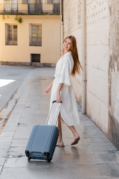 joven mujer alegre en vestido caminando con maleta en calle en valencia  - Foto, imagen