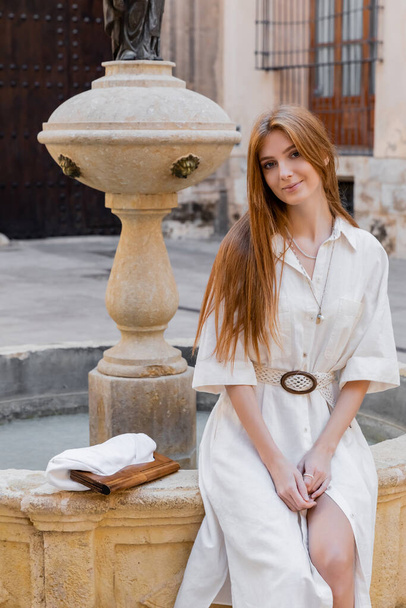 バレンシアの通りの噴水の近くに座っている白いドレスを着た笑顔の赤毛の女性 - 写真・画像
