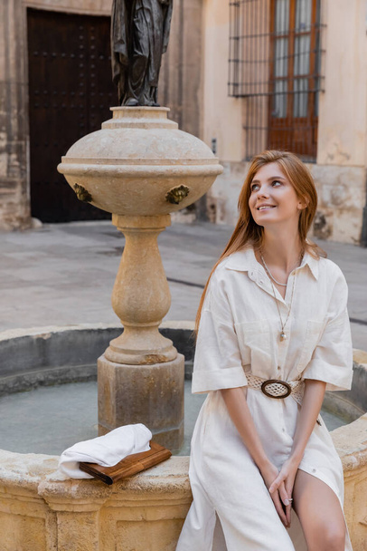 pleased woman in white dress sitting near fountain in valencia - Foto, afbeelding