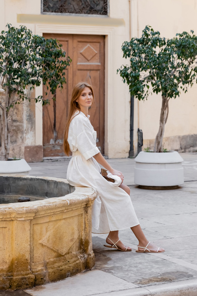 redhead woman in white dress sitting with purse near fountain on street of valencia - Foto, Imagen