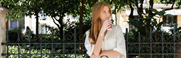 joven pelirroja en vestido sosteniendo café para ir cerca de valla en el parque verde europeo, pancarta - Foto, imagen