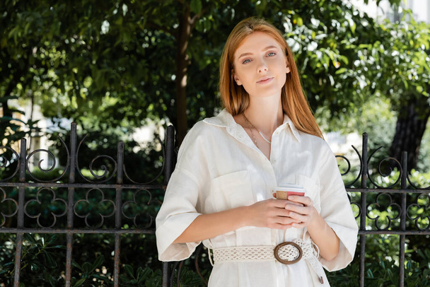 pelirroja en vestido blanco sosteniendo café para ir y mirando a la cámara cerca de valla en el parque verde europeo - Foto, imagen